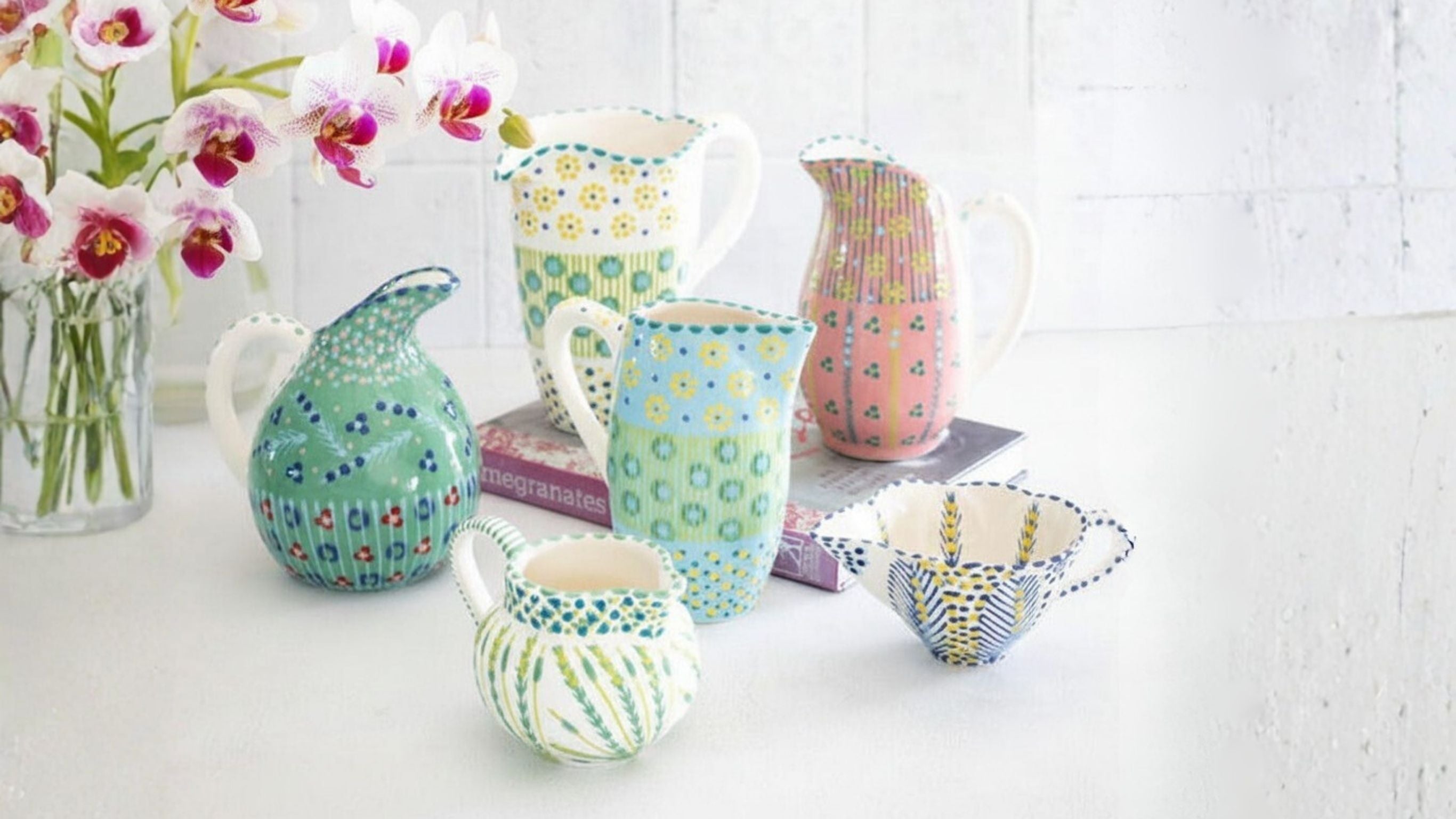 Colourful patterned tea and dessert serveware with jugs, cups, bowls, books, and flowers.