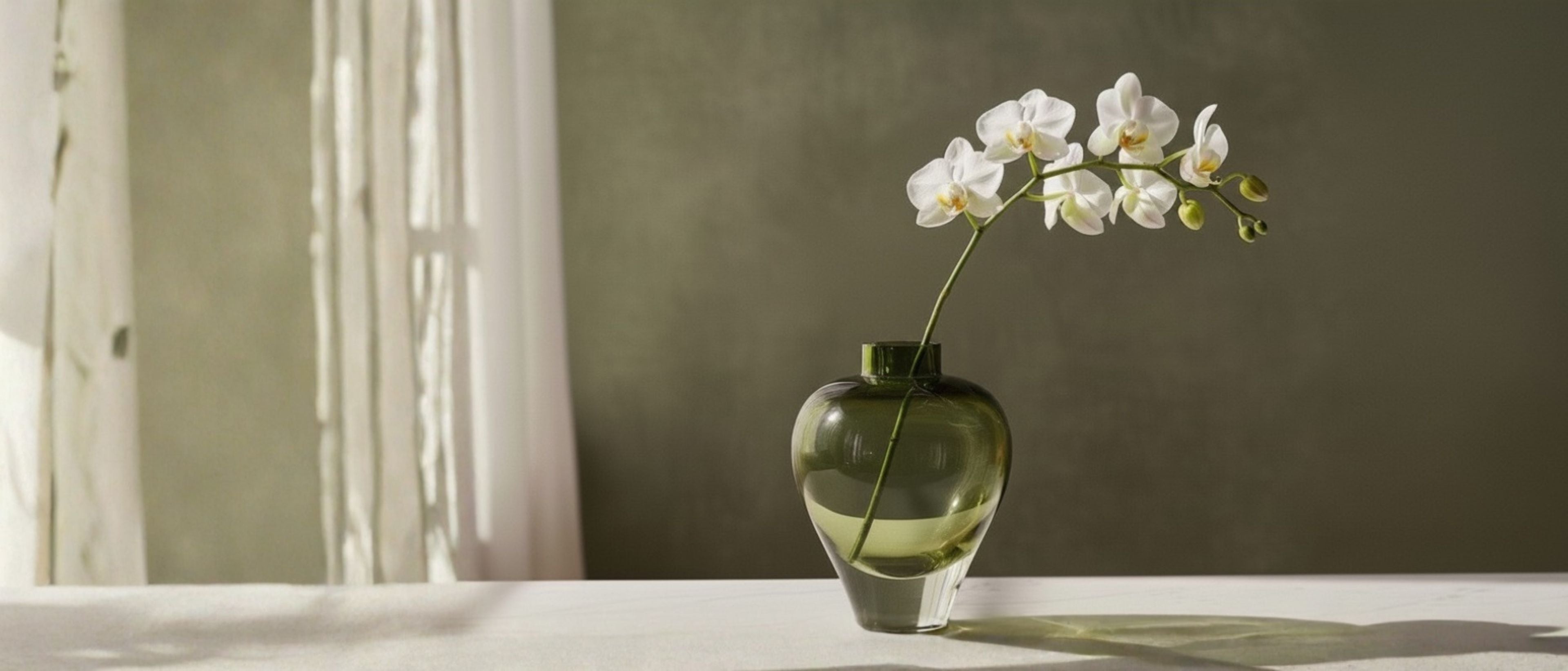 Green glass vase with white orchid flowers on a light surface.